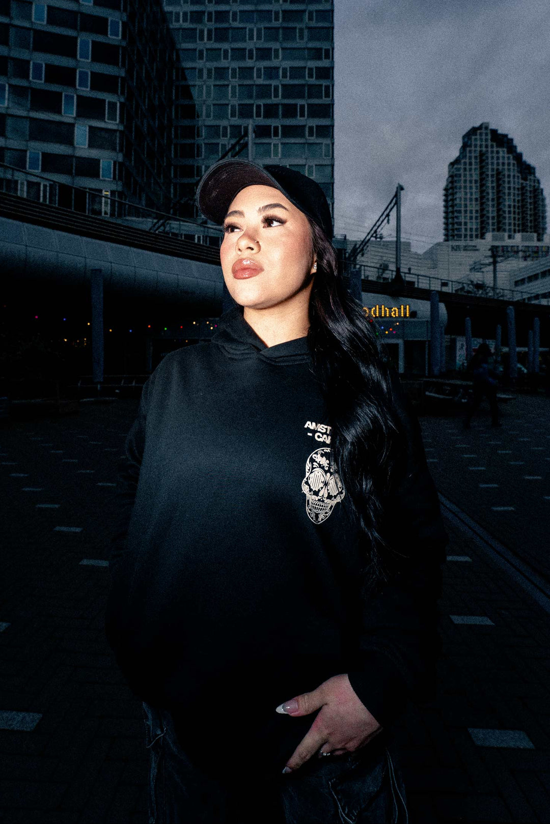 Person wearing a black hoodie and cap in an urban setting with buildings in the background